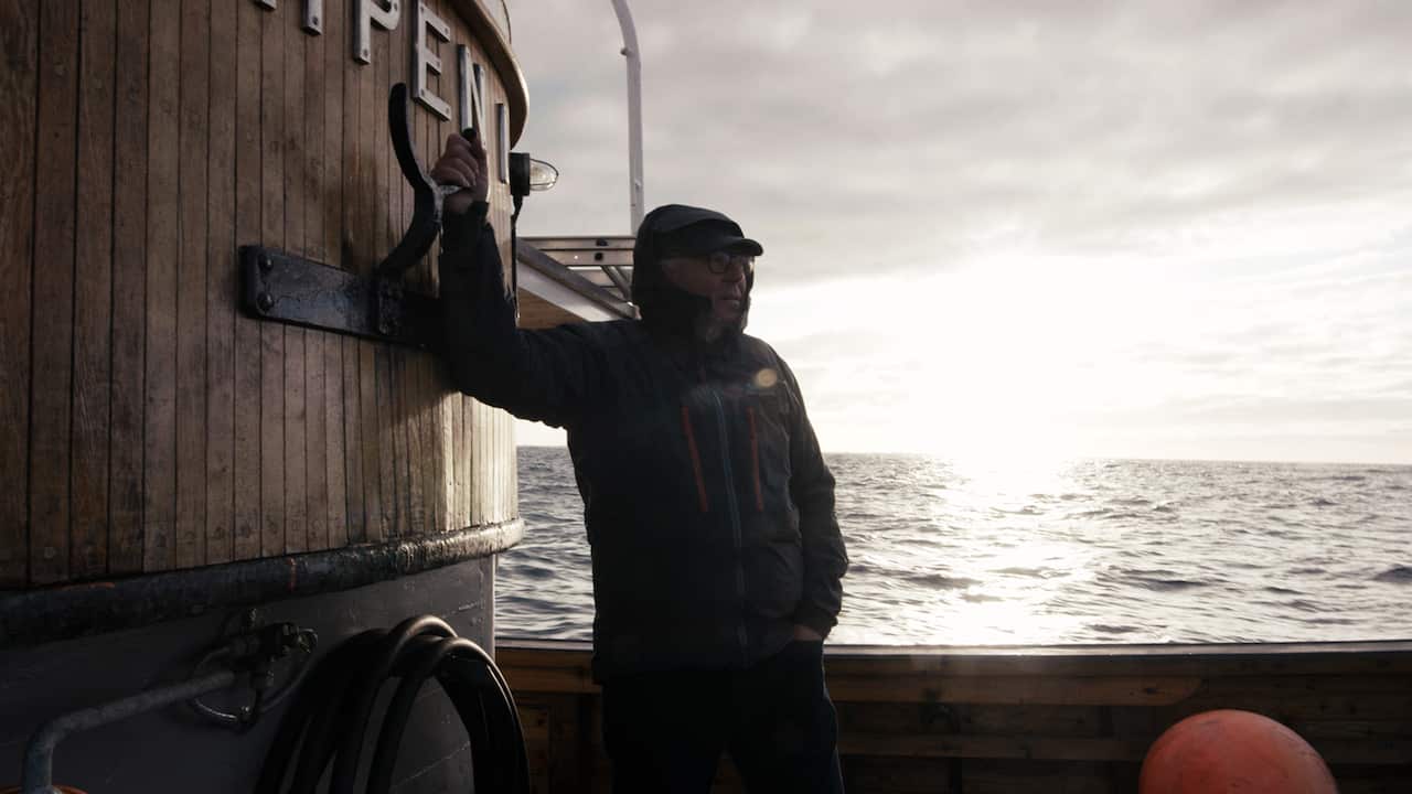 A man in a dark jacket stands on a boat, with waves stretching to the horizon behind him. 