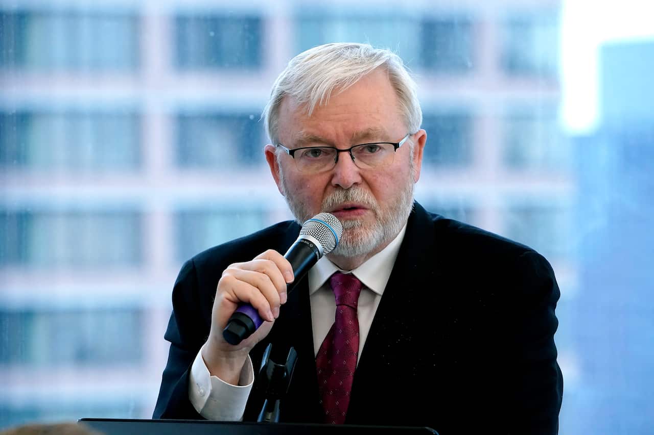A man wearing a black suit and a red tie is speaking into a microphone.