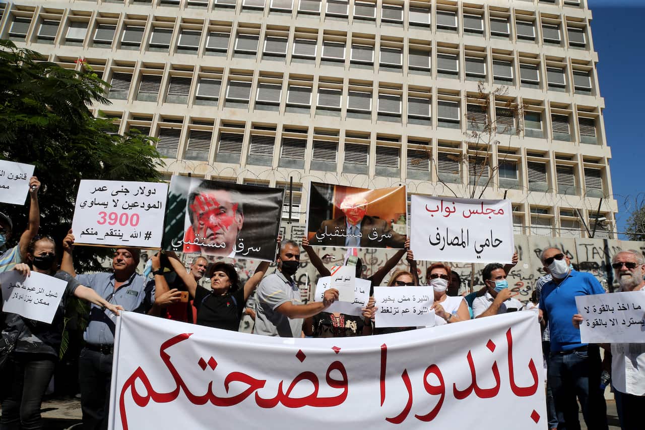 A group of people, some wearing medical masks, hold placards in Arabic at a protest outside of a government building.