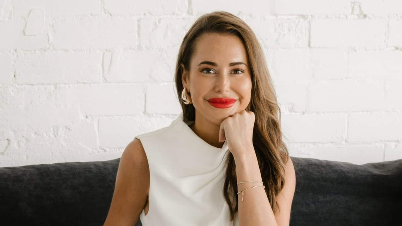 A woman with long, wavy brown hair in red lipstick and a white sleeveless top smiles on a couch with her hand resting on her chin