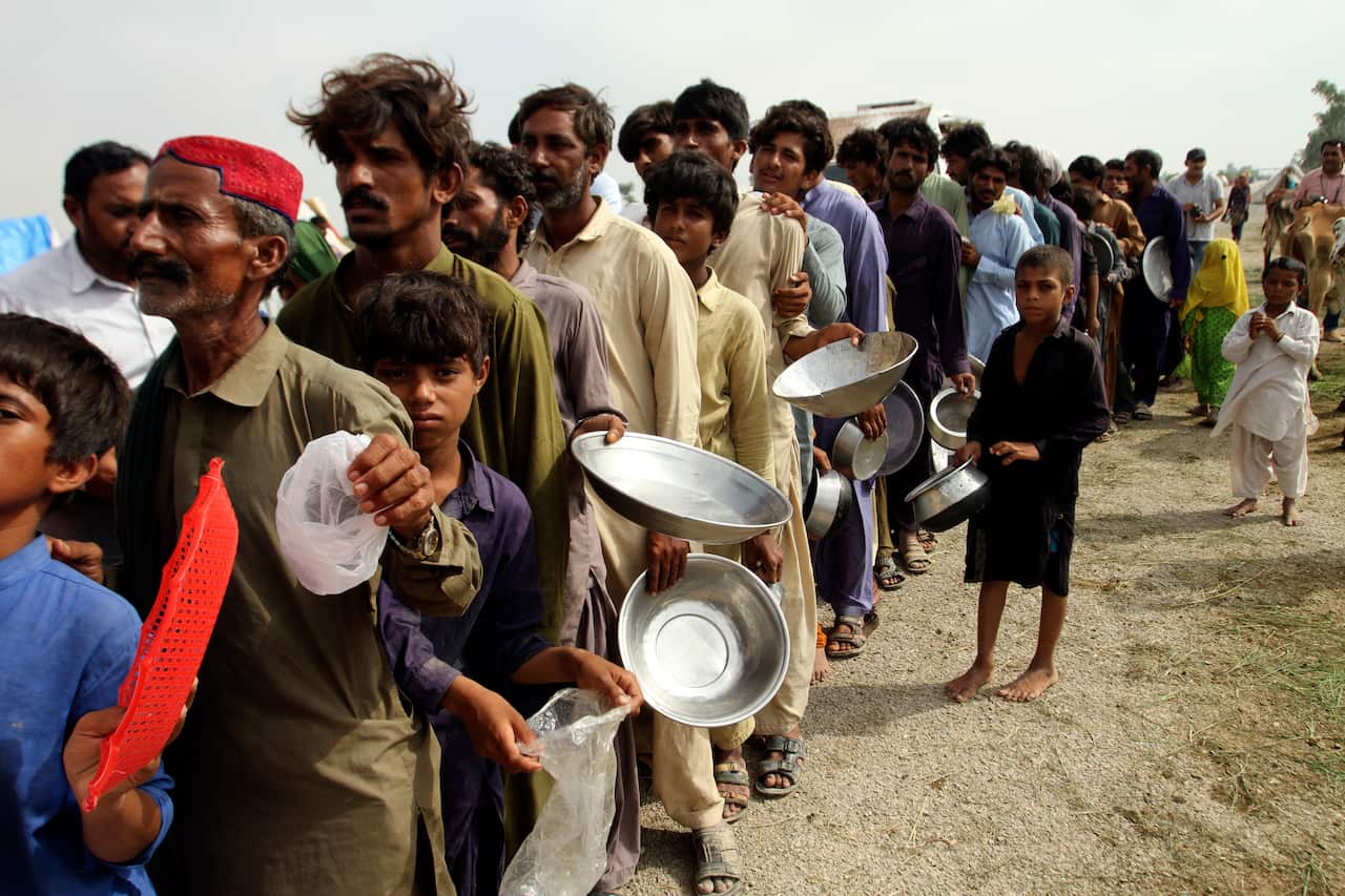 Flood affected people stand in a long line with utensils to get food.
