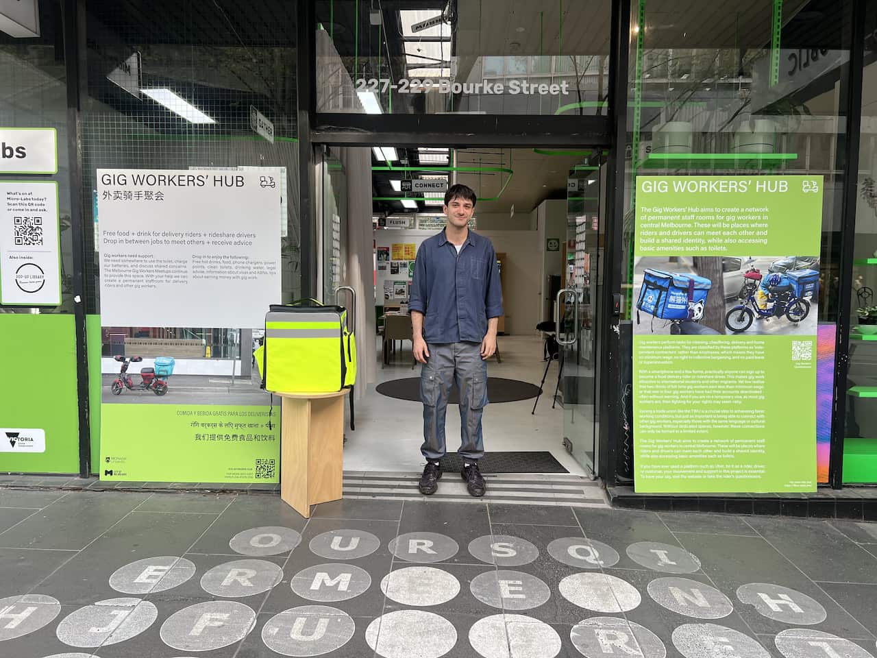 A man stands in the doorway of a shop front. There are signs on the front windows reading Gig Workers' Hub