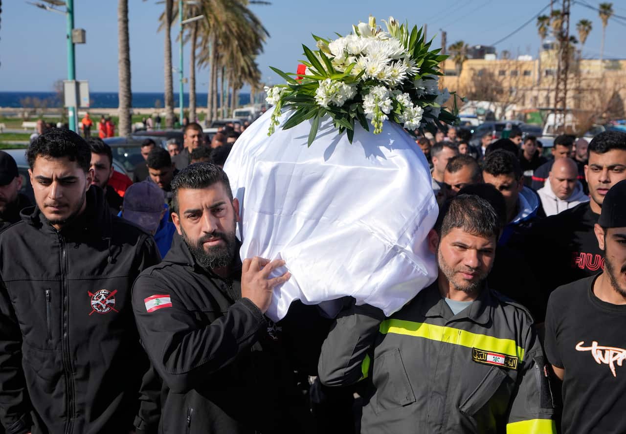 A group of men in black uniforms carry a coffin adorned with a wreath of white flowers during a funeral procession.