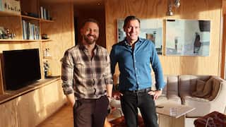 Two men smile for a photo standing in a lounge room. The man on the left has a beard and is wearing a brown checkered shirt and pants. The man on the right is wearing a blue shirt and black pants