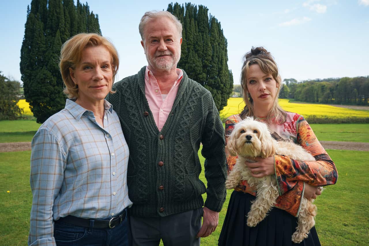 Three people stand together in a garden. A middle aged man and woman are beside a younger women, who is holding a fluffy dog. 