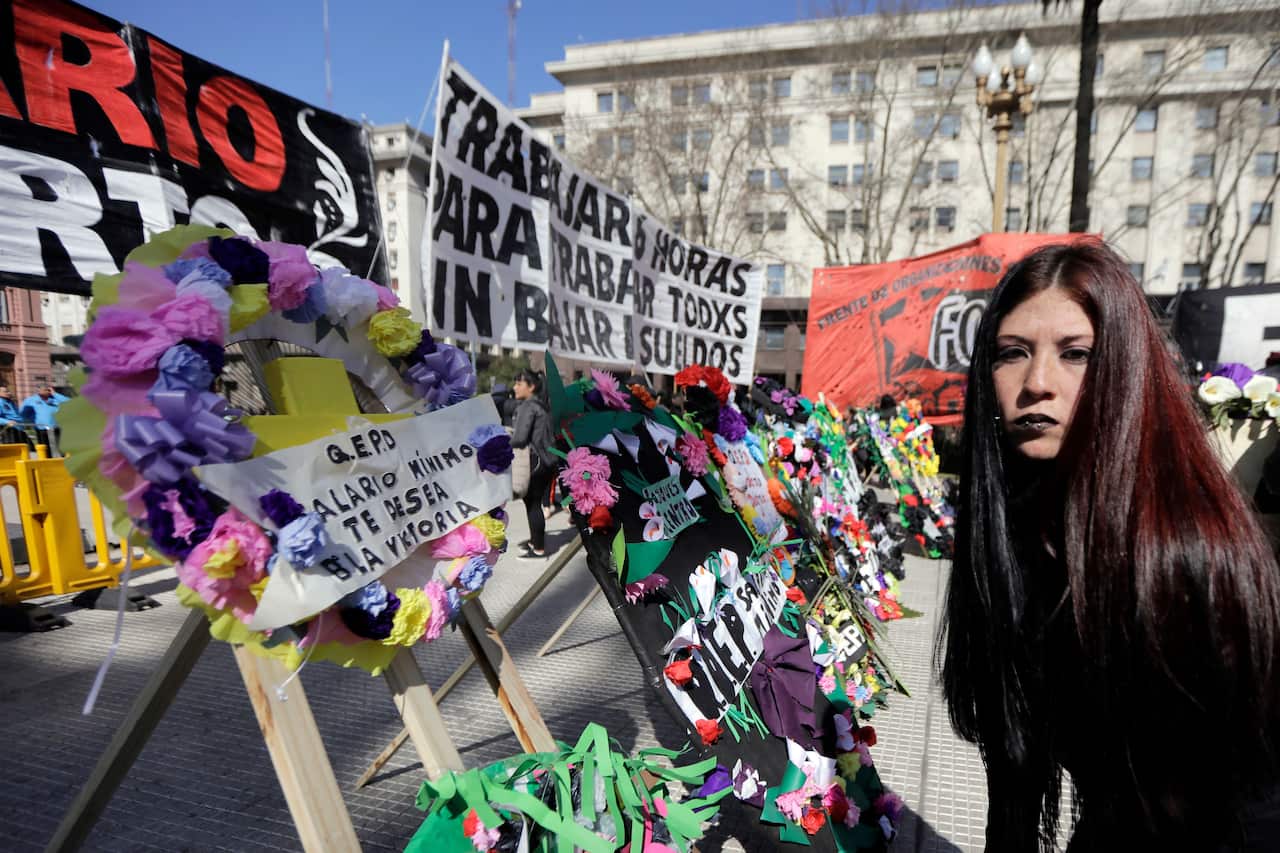An Argentine woman with dark hair and black lipstick stands in a public square surrounded by wreaths and signs