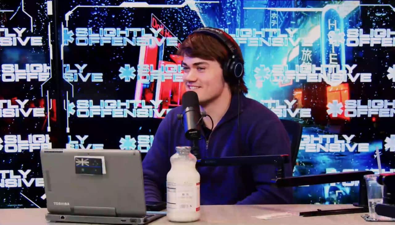 A man in a blue shirt, wearing headphones, is sitting at a table in front of a laptop. He is smiling as he speaks into a microphone in front of a well-lit background with the words "Blightly Offensive" written on it several times.