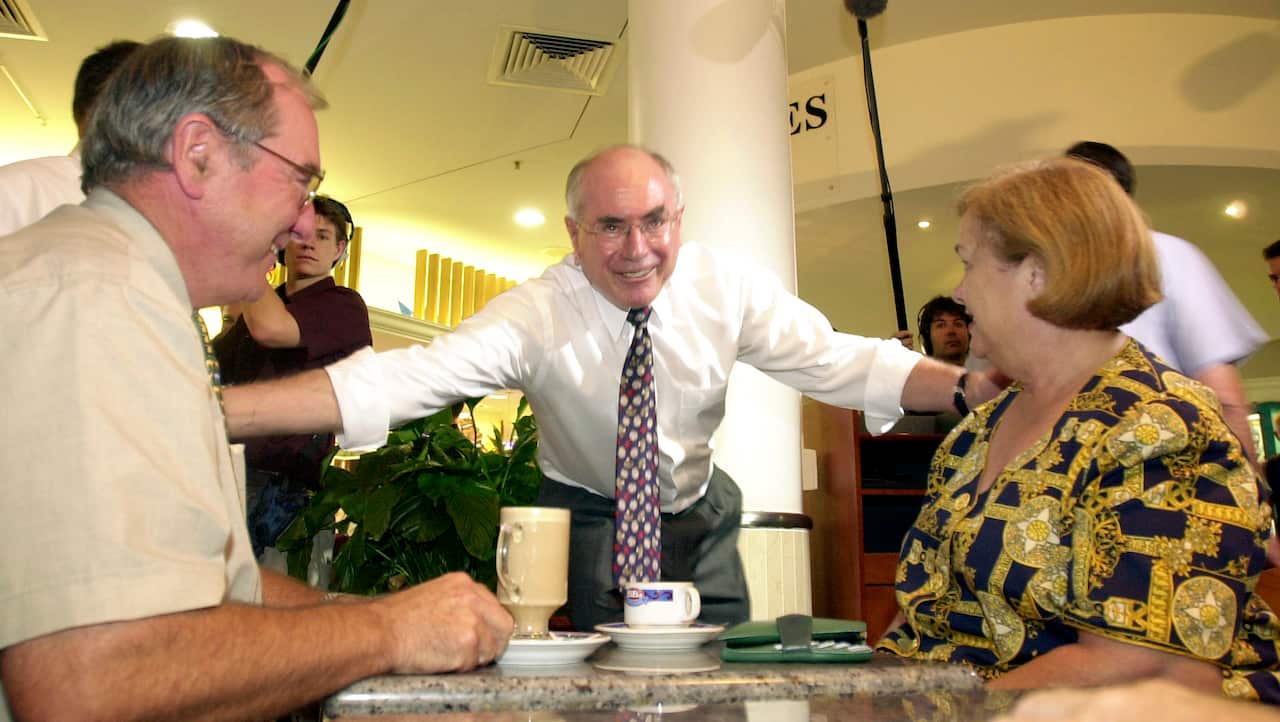 John Howard crouched, with one arm each on a man and a woman seated at a table drinking coffee. There are media personnel behind them.
