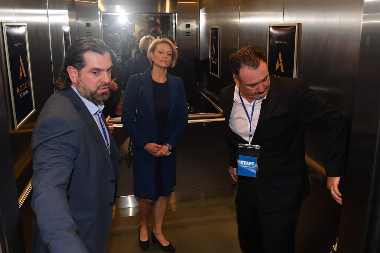 A blonde woman stands in a lift between two brunette men