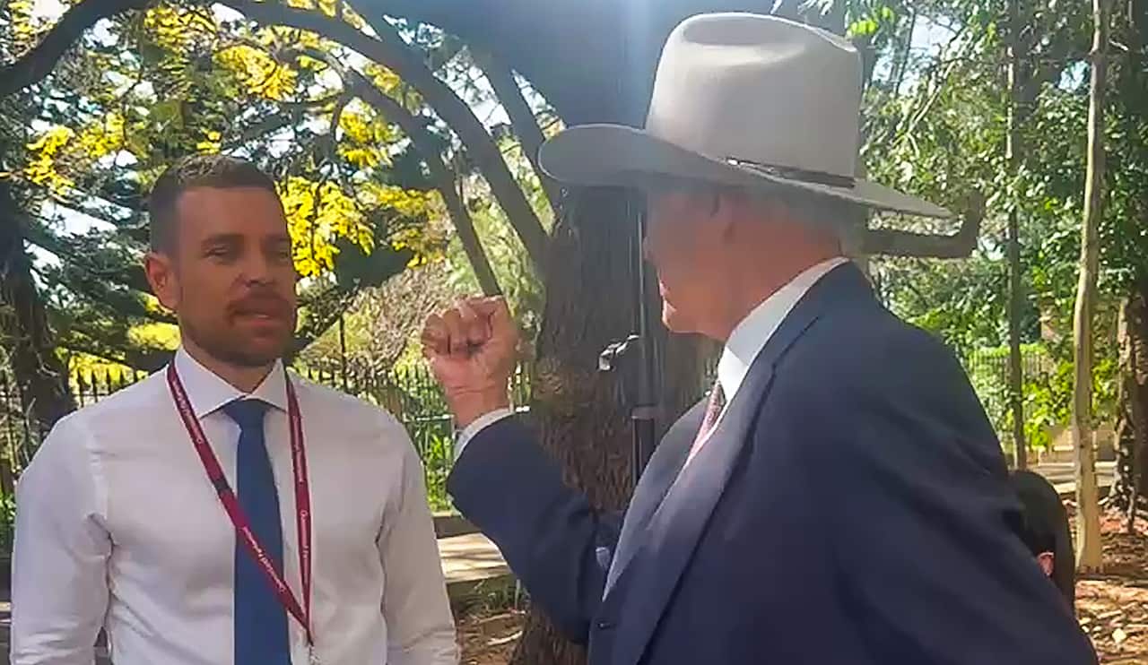 A medium close-up of a man in a blue suit and white cowboy hat speaking passionately, his right hand clenched in a fist. He is facing another man in a white dress shirt, a blue tie, and a red lanyard. They are standing outdoors, with trees and sunlight in the background.