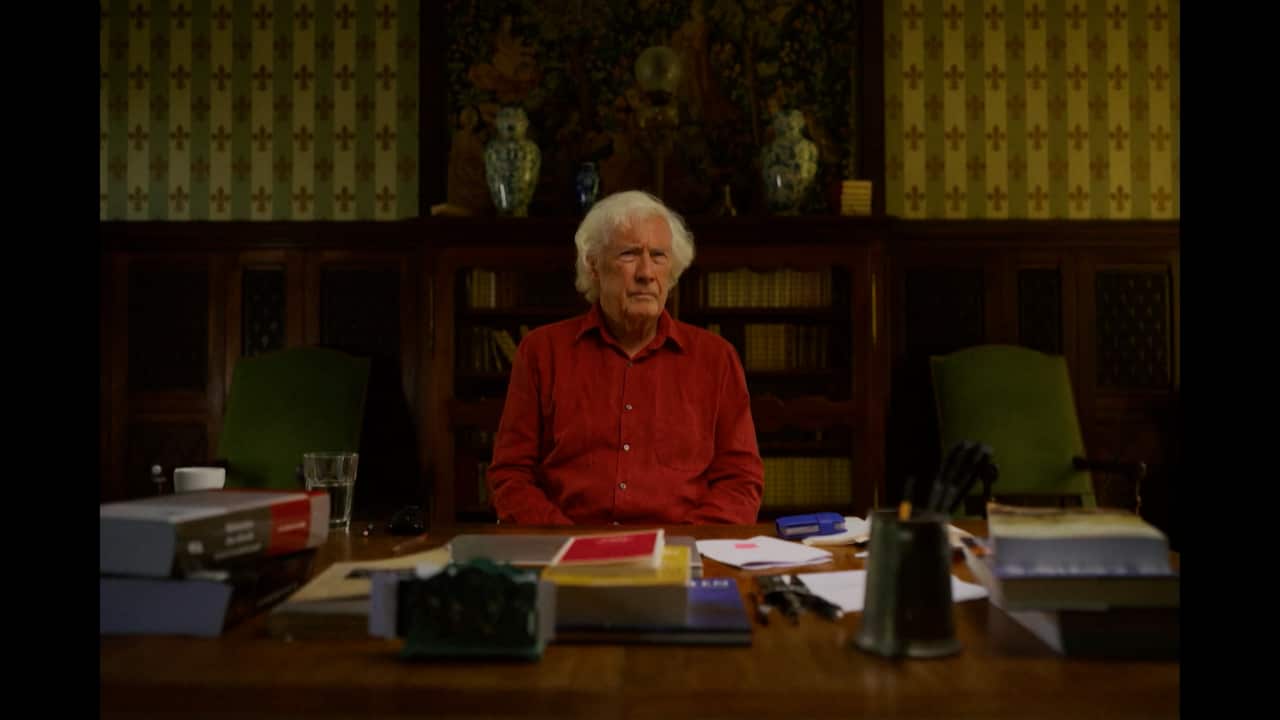 A grey-haired man in a red shirt sits at a desk covered with books and papers in a study.