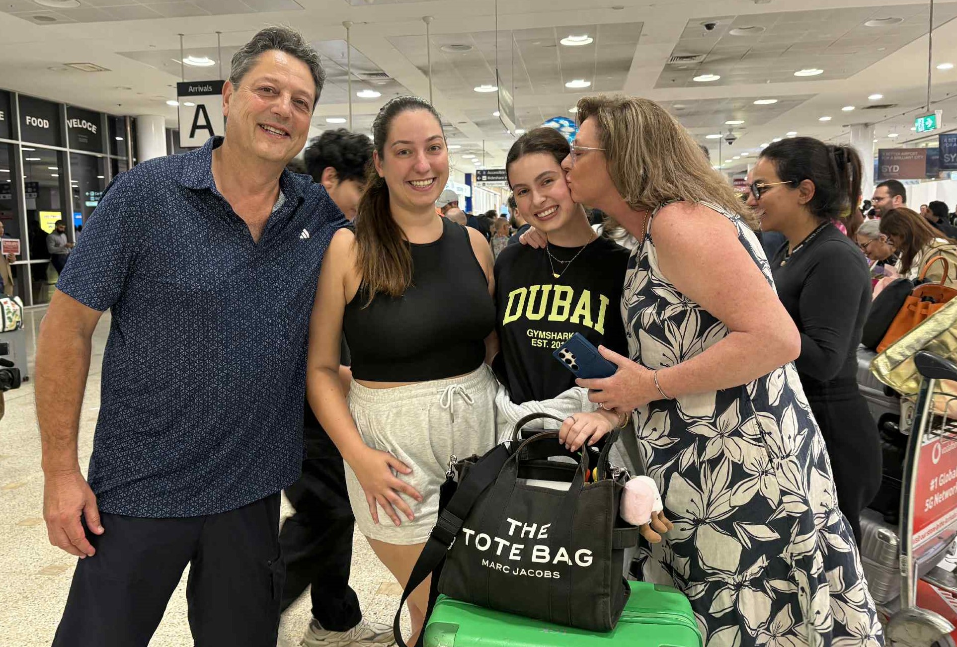 Four people standing in a row, from the left, a middle aged man in dark navy shirt, a young woman in dark hair wearing a black tank top and grey gym shorts, a young woman smiling with dark hair wearing a black top with DUBAI written on it in yellow block letters, and next to her a middle aged woman kissing her.
