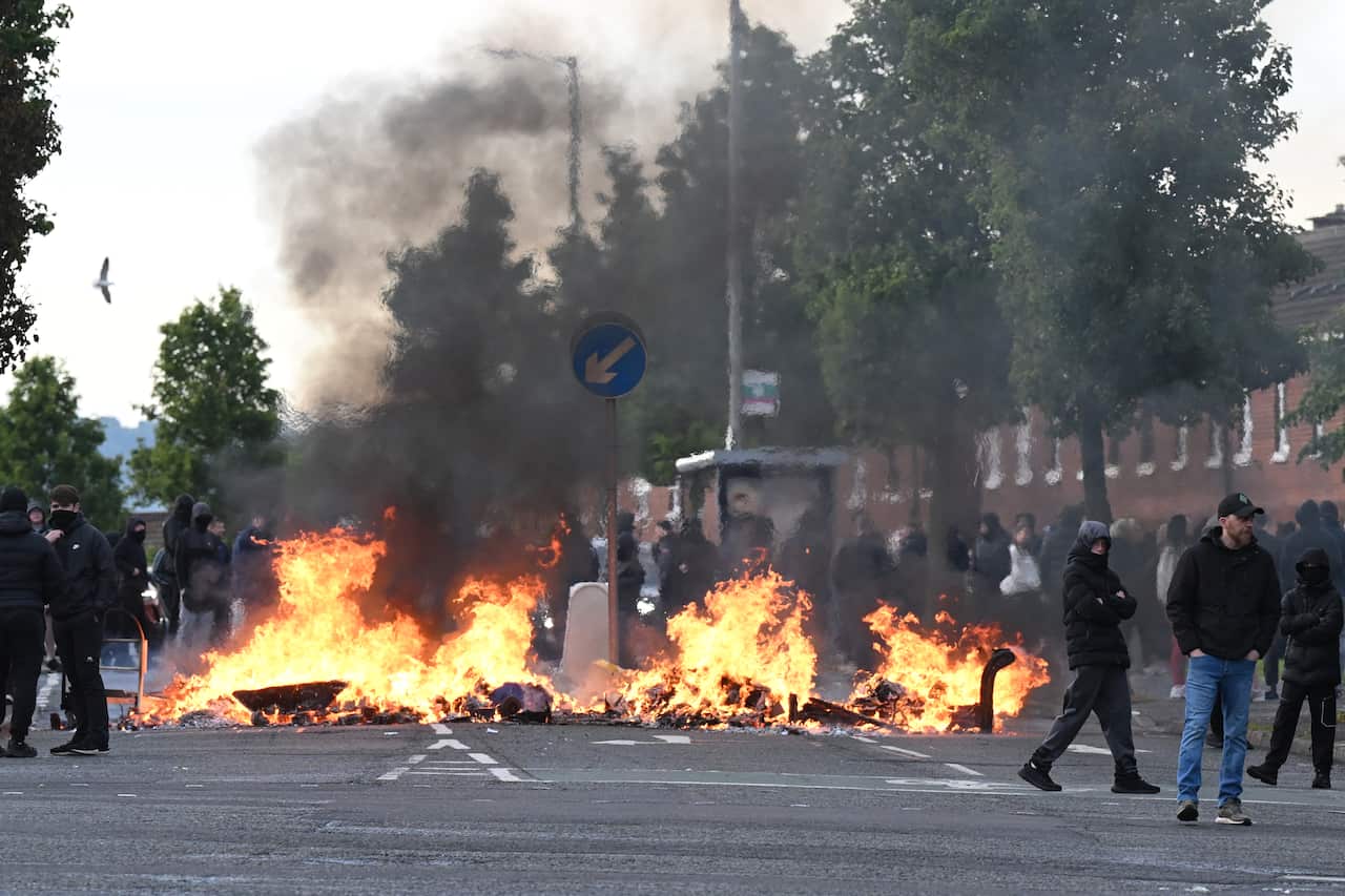 People stand near burning debris and smoke in Belfast during protests following a stabbing incident.