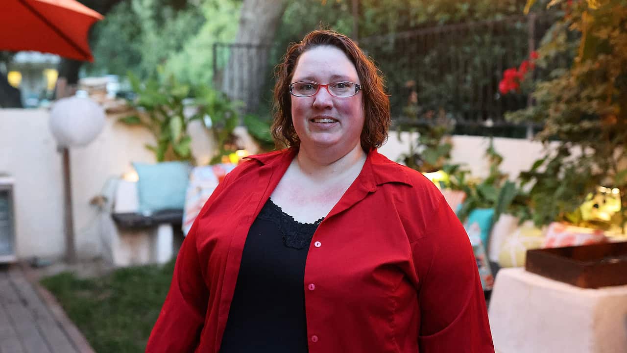 A red-haired woman wearing reading glasses and a red jacket over a black top 