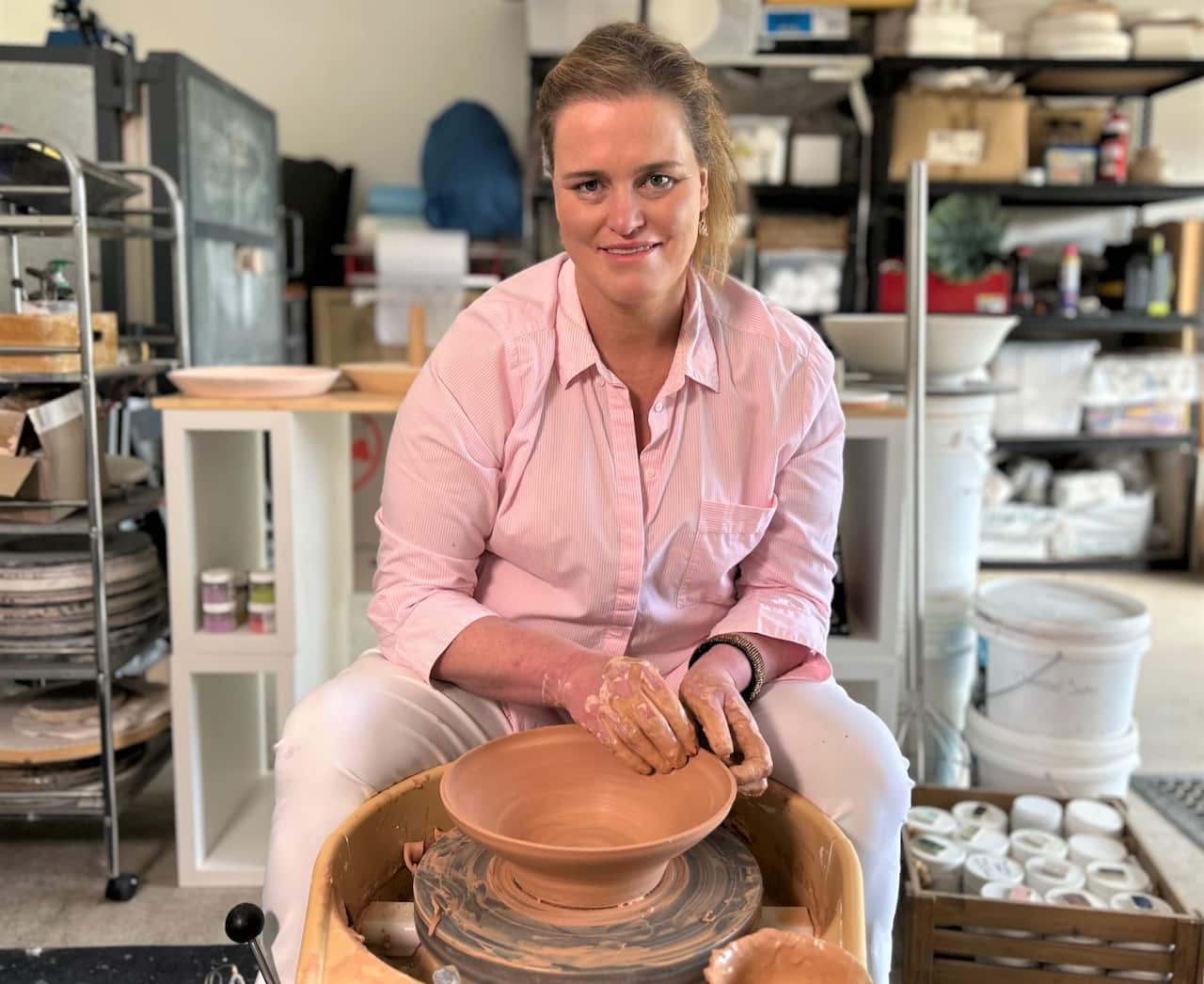 A woman wearing a pink shirt sits at a pottery wheel moulding a pot.