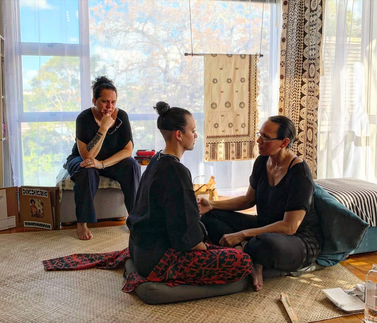 Two women sit opposite each other on the floor as another looks on