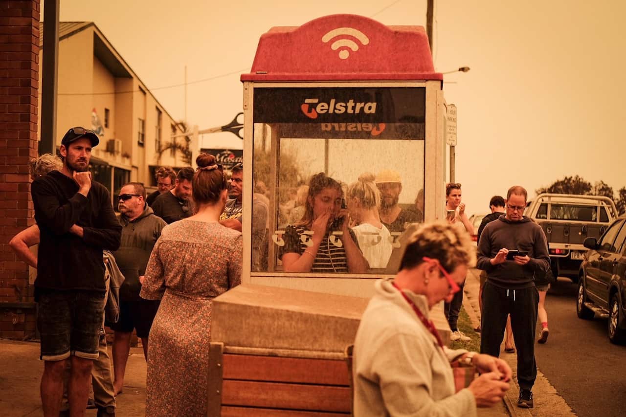 People gather around a Telstra public phone booth during smoky conditions, with several individuals using mobile phones or looking at devices along a street.