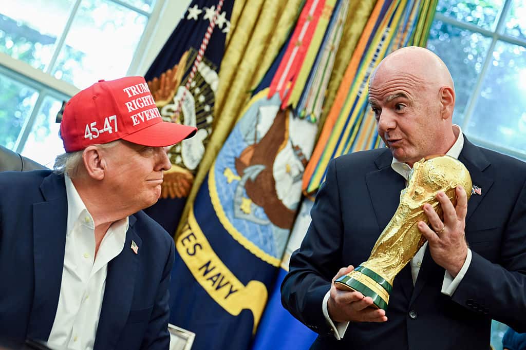 Donald Trump, wearing a red cap reading "Trump was right about everything" sits next to Gianni Infantino, who is standing and holding a gold trophy