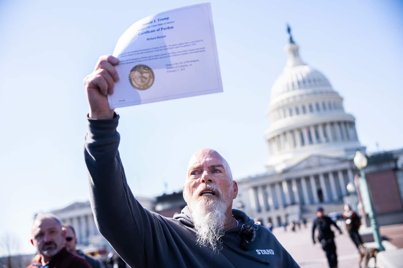 Middle aged man with bald head and white beard holds up a presidential pardon certificate in front of the US Capitol.