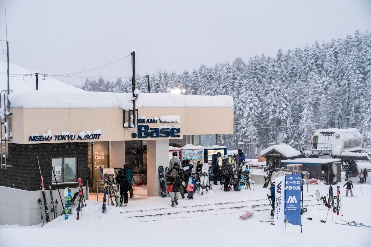 A building covered in snow with people wearing ski gear standing outside. Snowboards and skis line the building. 