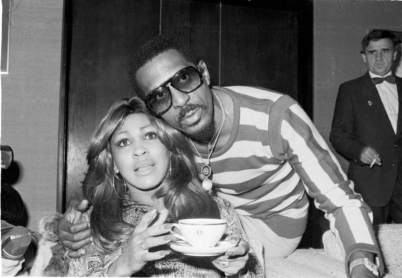 Black and white photograph of Tina Turner holding a cup of tea with former husband Ike Turner's arm around her