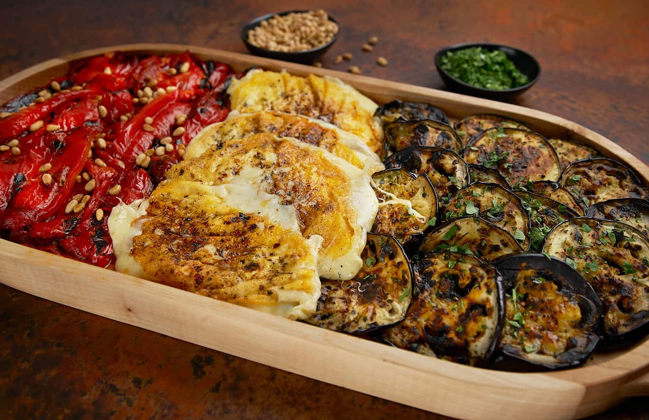 Grilled capsicum, cheese and eggplant sit in a long wooden platter. The cheese has a lovely golden brown crust, the capsicum is topped with scattered pinenuts and there is chopped herb on the eggplant slices. 