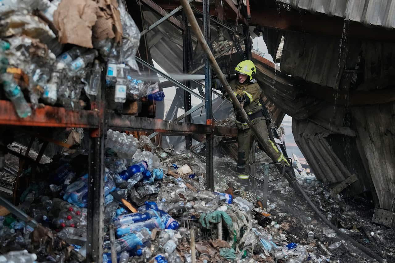 A firefighter wearing a bright yellow helmet blasts water at a warehouse filled with plastic bottles, destroyed by a fire.