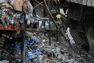 A firefighter wearing a bright yellow helmet blasts water at a warehouse filled with plastic bottles, destroyed by a fire.