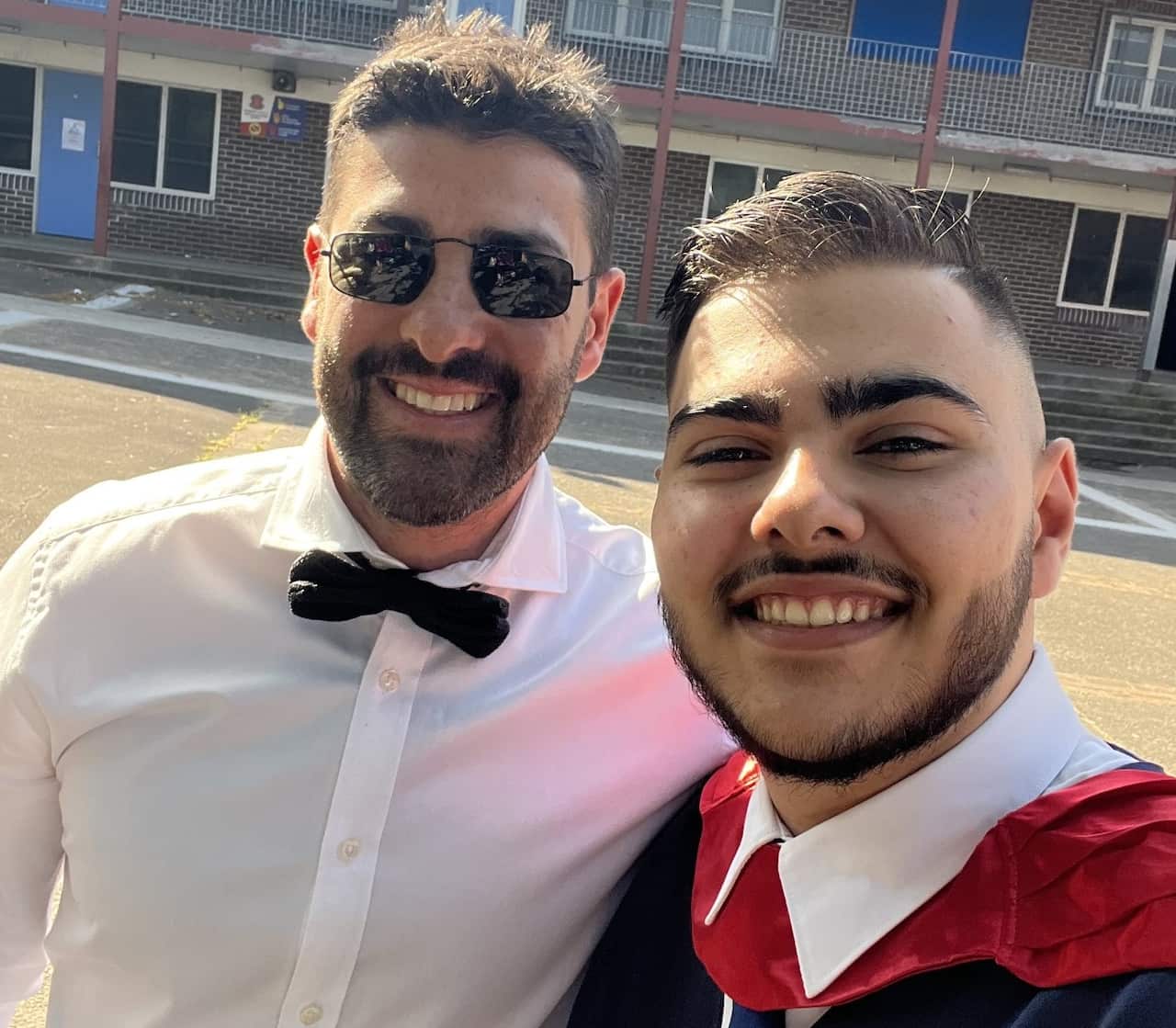 Two men, one in a white shirt and the second one in graduation regalia are smiling. 