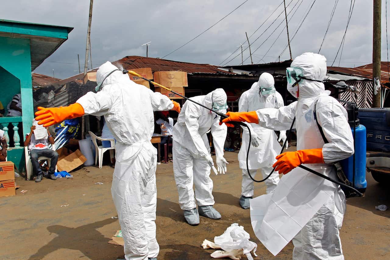 Health workers in white suits standing on a dirt path. One is washing another with a high-powered hose.