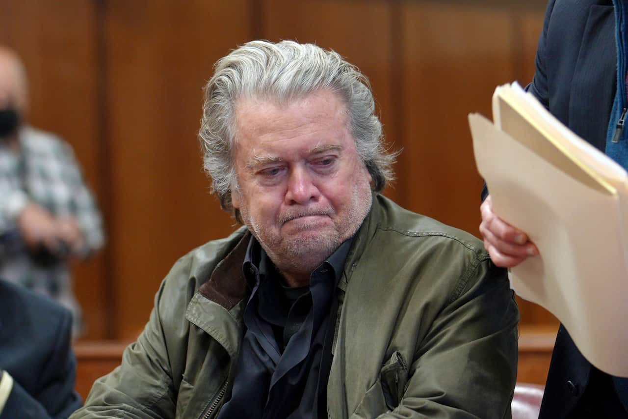 Steve Bannon sitting in a courtroom. Someone next to him is holding a folder with documents in it.