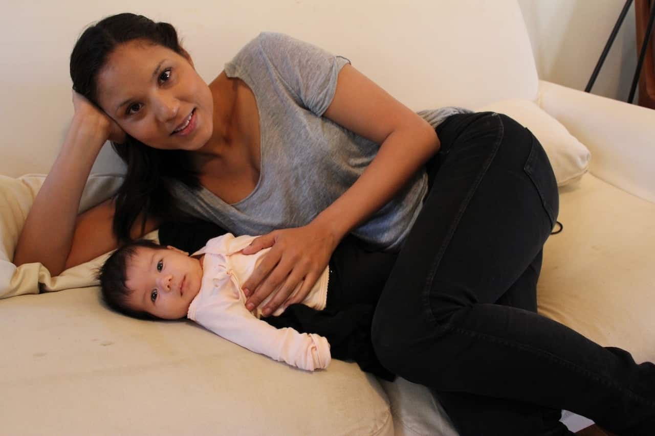 A woman in a grey t-shirt and black pants is lying on a bed and looking into the camera as she's put her hand on an infant's belly 