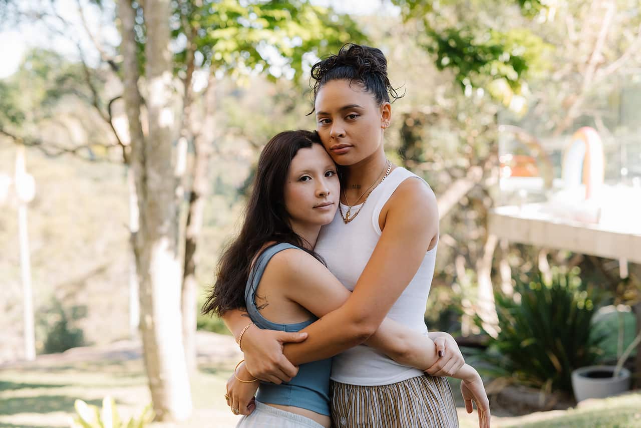 Two people stand with their arms around each other. They are standing in front of a light, sunny garden. 