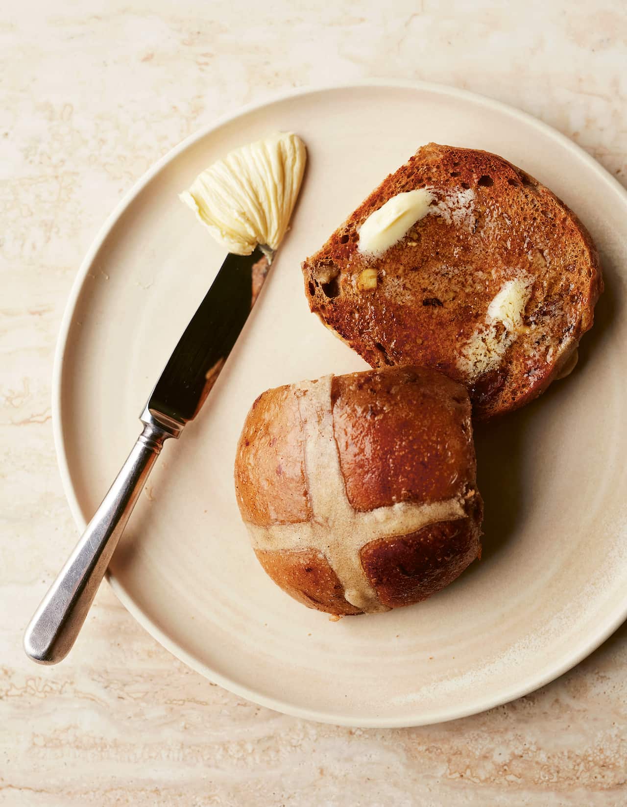 A split hot cross bun sits on a plate, the bottom has a pale spread melting on it. A knife wiht more spread sits alongside. 