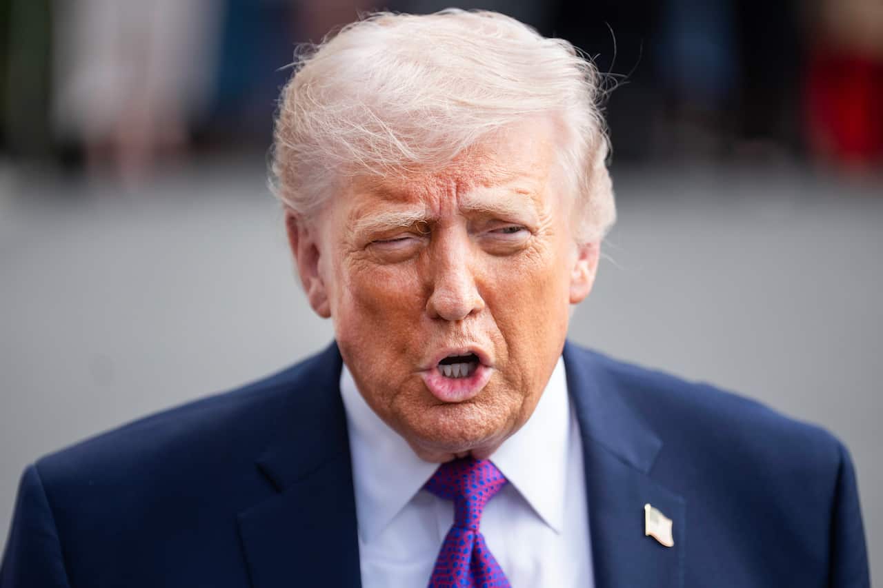 Donald Trump, wearing a blue blazer, white shirt, purple and blue tie, and American flag pin.