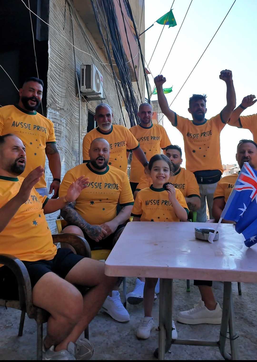 A young girl sits at an outside table surrounded by cheering men. They're all wearing green and gold Aussie Pride T-shirts.