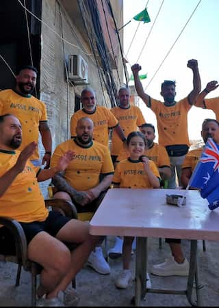 A young girl sits at an outside table surrounded by cheering men. They're all wearing green and gold Aussie Pride T-shirts.