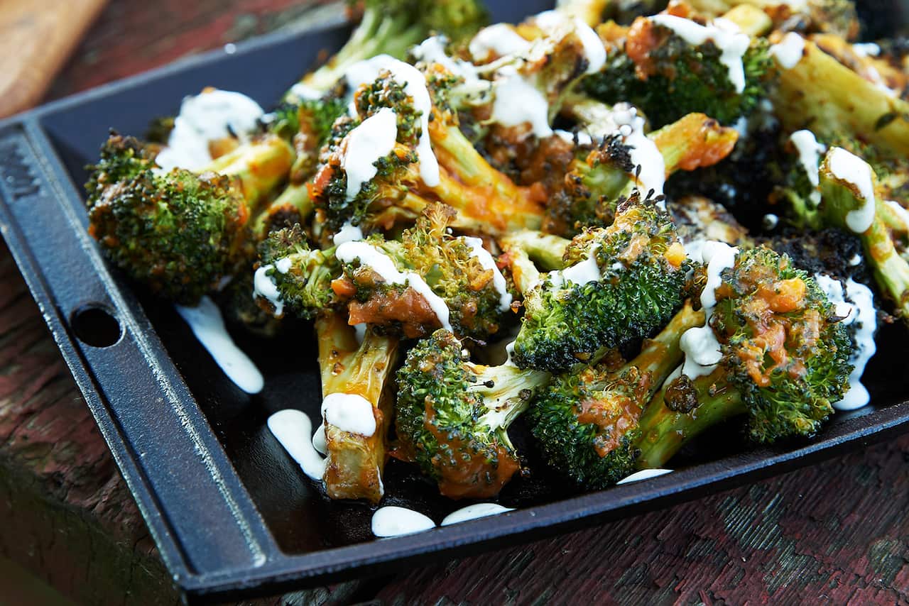 Cooked broccoli florets sit in a cast iron tray, drizzled with a brown sauce and a creamy sauce.  