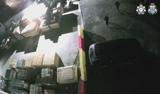 An aerial view of a warehouse. A car and two people clad in black clothing are on the ground.