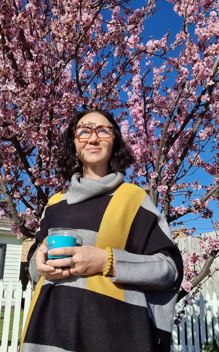 A woman wearing a jumper stands with a cup in her hand. Behind her there is blue sky and a cherry blossom tree.