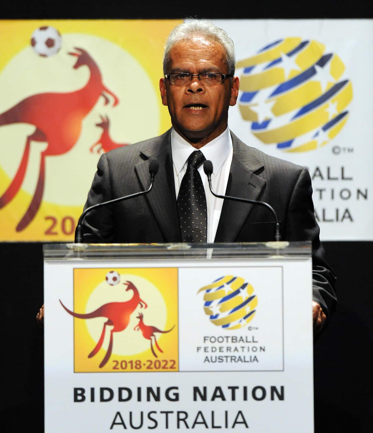 harry williams wearing spectacles stands speaking at a lectern 