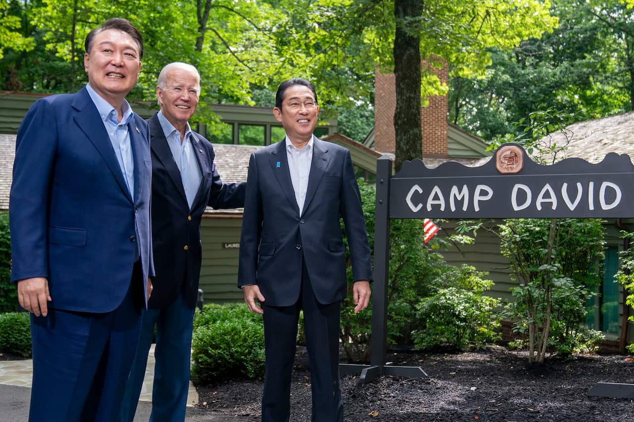 Three men wearing suits standing next to a sign that reads "Camp David".