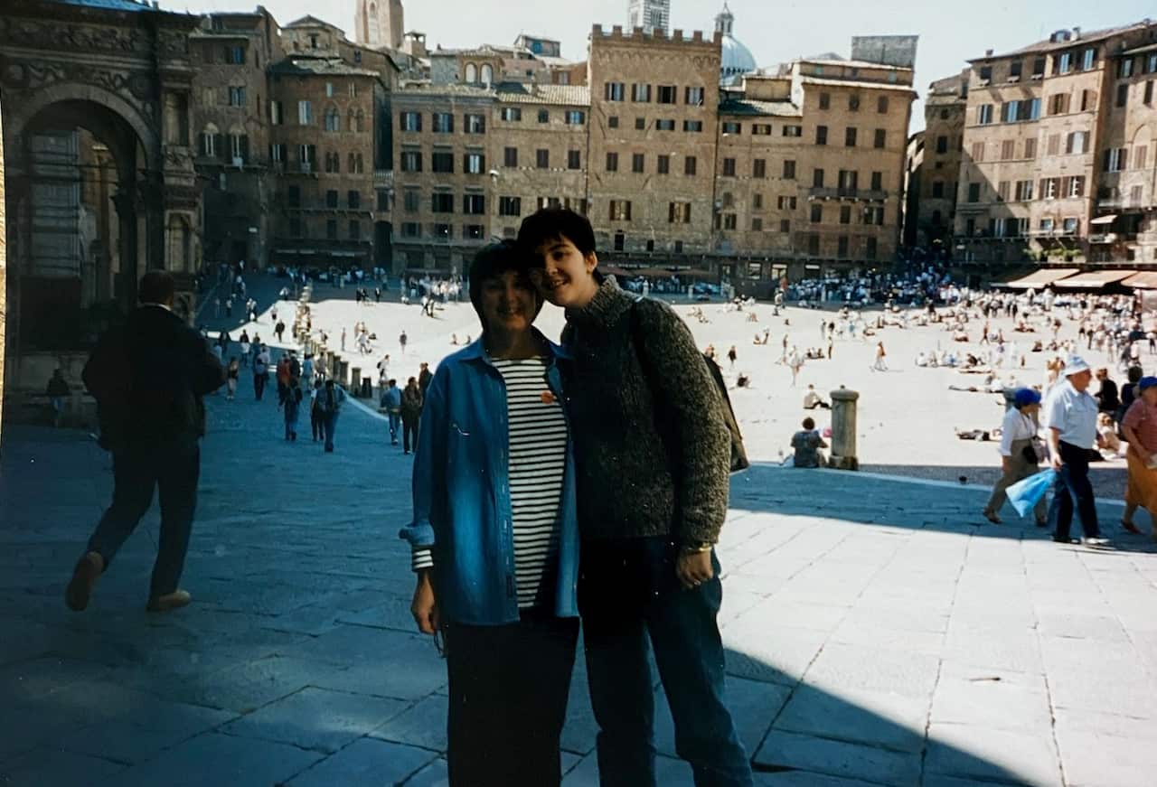 A mother (left) and daughter stand together and smile on a sunny day in a town square somewhere in Europe.