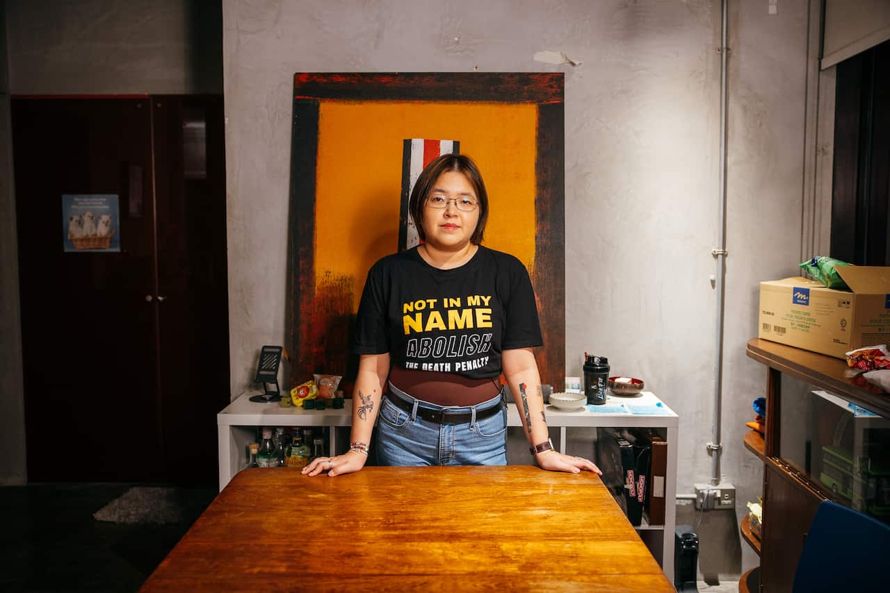 An Asian woman with short hair and glasses stands in an office room, resting both hands on a table. She is wearing a black T-shirt with the text 'Not in my name. Abolish the death penalty' printed on it
