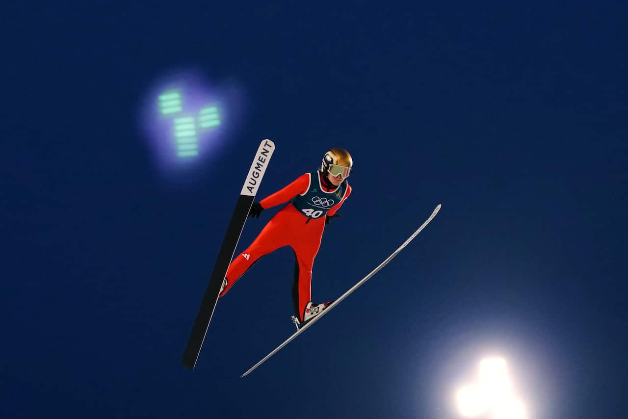 A woman wears a red jumpsuit, a helmet and goggles while skiing. She soars high in the sky during a jump.