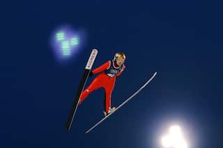 A woman wears a red jumpsuit, a helmet and goggles while skiing. She soars high in the sky during a jump.