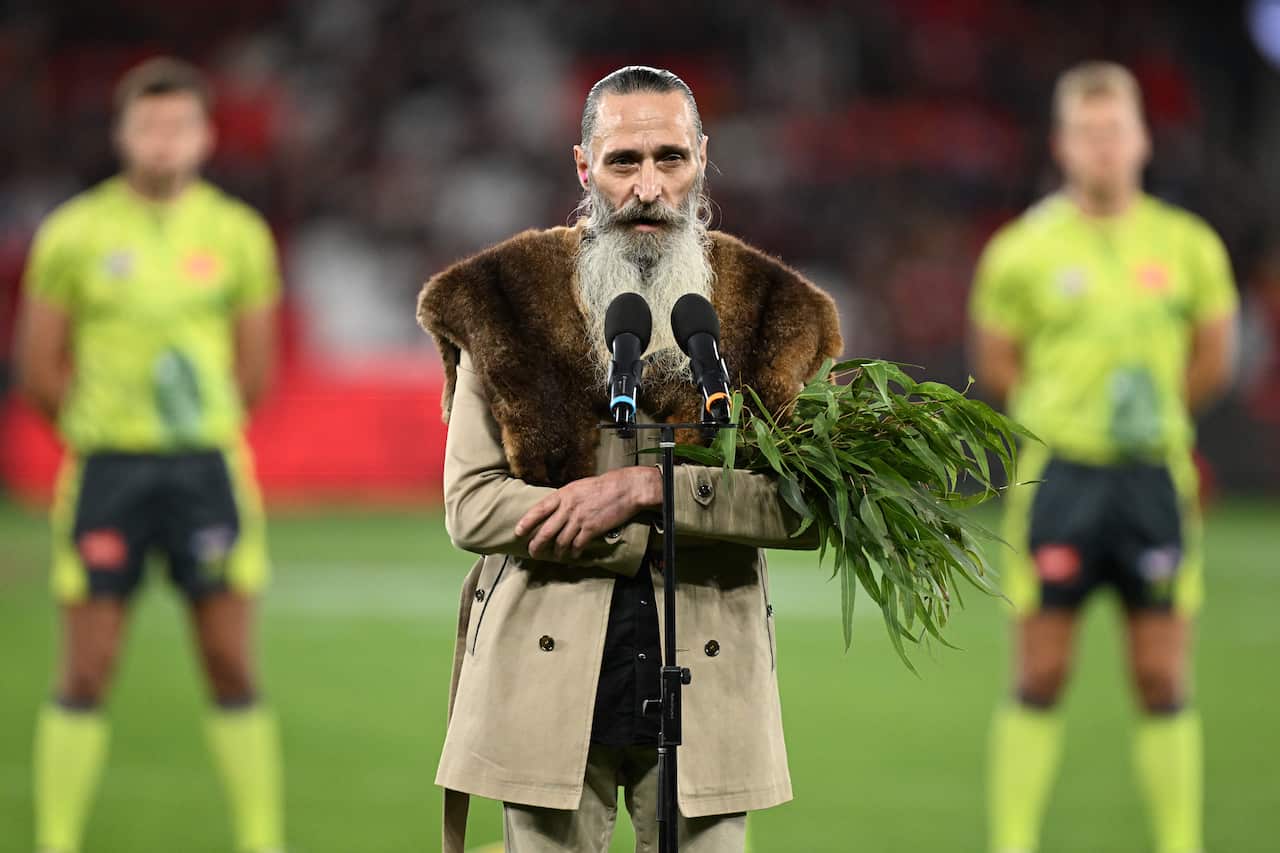 A man wearing a jacket with a fur over his shoulders and carrying a sheaf of gum leaves stands behind a microphone on a sporting field