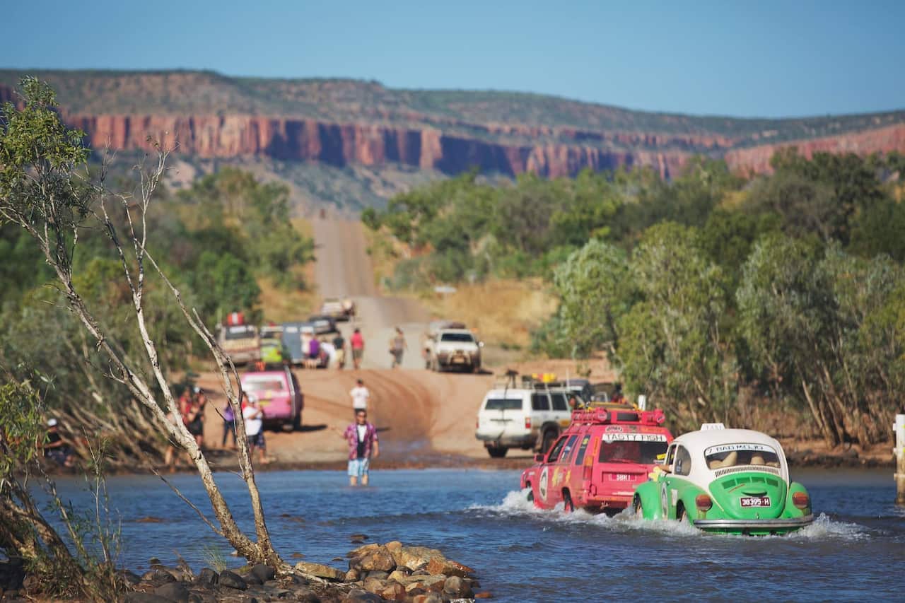 Colourful small cars drive through a river with a backdrop of the Kimberley mountains.
