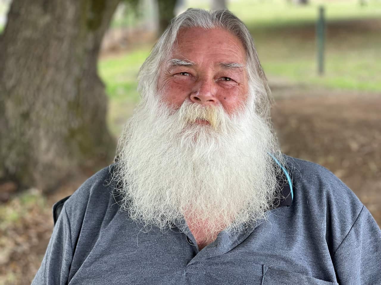 Kamilaroi man Mitchum Neave looks directly to camera. He has a long white beard and moustache, and smiling eyes. He is wearing a grey/blue t-shirt. A head and shoulder shot.