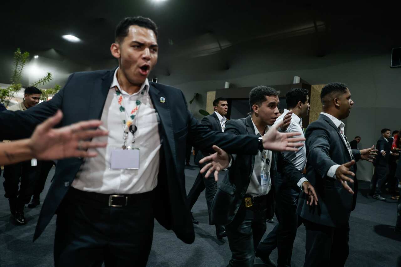 A row of men in suits and lanyards hold their arms out as they move through a large, dark indoor space.
